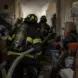 Firefighters advancing a hoseline through a cluttered attic toward a concealed knee-wall fire.